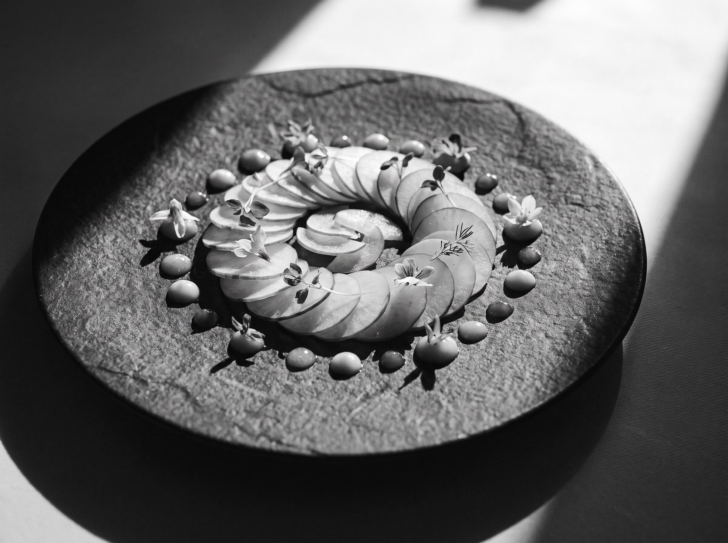 Hauchdünne Scheiben in einer Spirale auf dunklem Steinteller, mit Saucenpunkten und Kräuterspitzen angerichtet, Schwarzweiss-Aufnahme